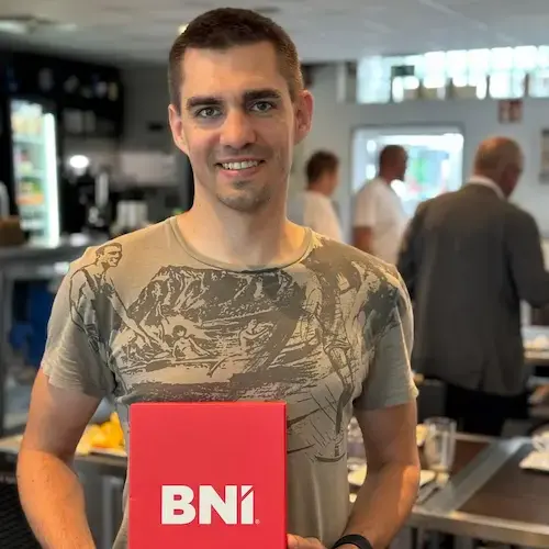 Lächelnder Mann mit kurzen Haaren in olivgrünem T-Shirt hält eine rote BNI-Mappe vor sich, im Hintergrund ein Café mit weiteren Teilnehmern eines Netzwerktreffens.